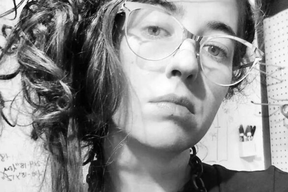 A black and white headshot of Amanda Fay. Her long medium colored hair is pulled into a messy updo. She wears clear glasses and a dark top. With her head tilted slightly to the side, she looks straight into the camera with a serious tone.