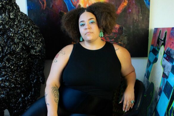 A color, full body portrait of Alexis Hunter. She sits in front of her colorful paintings. She wears a black sleeveless top and black pants and sits in a relaxed pose. She looks directly into the camera with a serious tone.