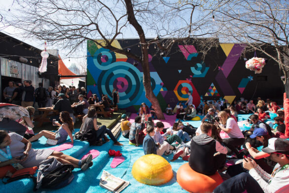 A photograph of a large crowd of people seated in an outdoor patio. The patio is covered in blue astro turf and people are seated on brightly colored round seats. There is a vibrant abstract mural on the exterior of a building behind the crowd.