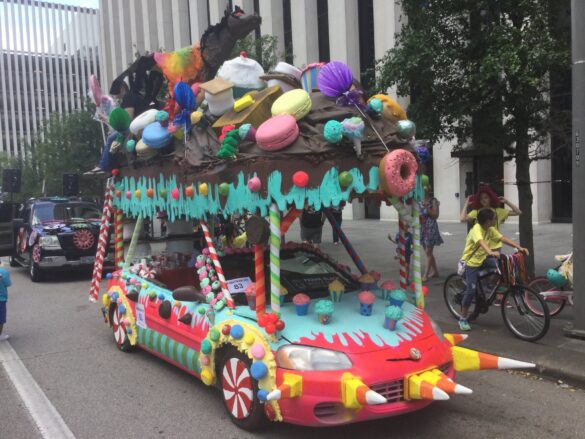 A photograph of a convertible red car transformed into an art car featuring an assortment of treats including sculptures of candy, ice cream, donuts, cupcakes, etc.