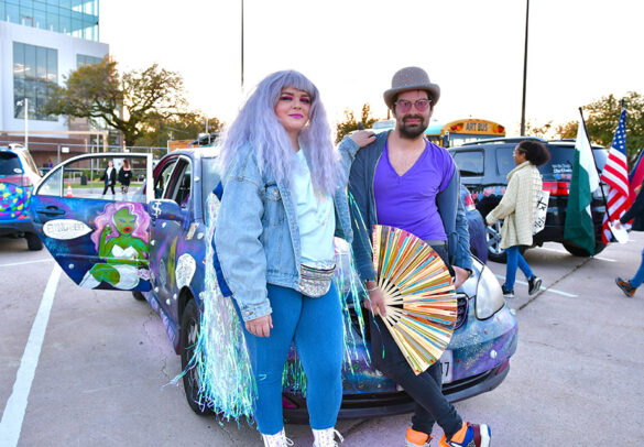 A woman and man wearing bright purples and blues stand in front of their art car at the 2022 Art Car Parade Kickoff.