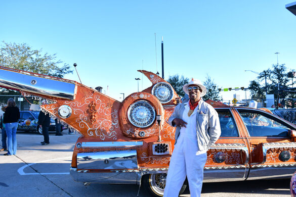 An artist wearing a painters jumpsuit, jacket, hat, and sunglasses poses in front of his orange art car at the 2022 Art Car Parade Kickoff.