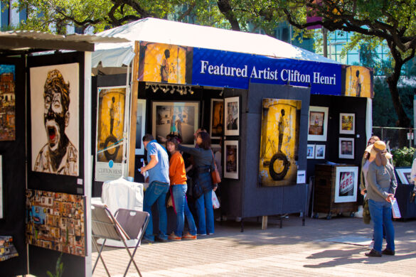 A photograph of an artist's booth at the Bayou City Art Festival. The booth displays an array of images and a small group of people are looks at the art on display.